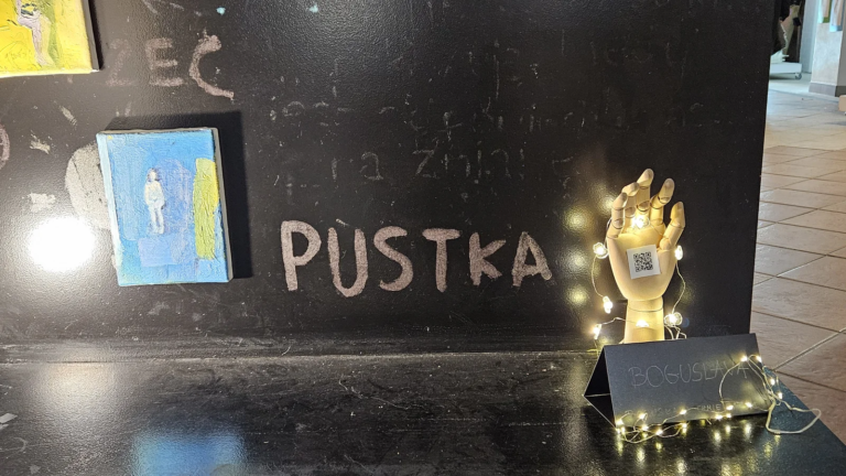 Miniatura z postacią, napis kredą Pustka i drewniana dłoń z kodem QR w lampkach
